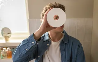 Ein Mann In Einem Badezimmer Blickt Durch Die Mitte Einer Klopapierrolle In Die Kamera.