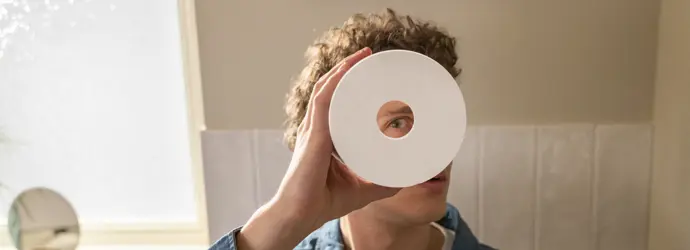 Ein Mann In Einem Badezimmer Blickt Durch Die Mitte Einer Klopapierrolle In Die Kamera.