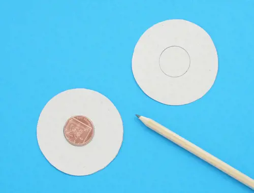 Два отрязани картонени кръга върху синя повърхност с монета в центъра и молив отстрани.