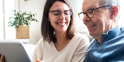 Eine Frau und ein älterer Mann lachen und teilen sich ein Tablet auf der Couch.