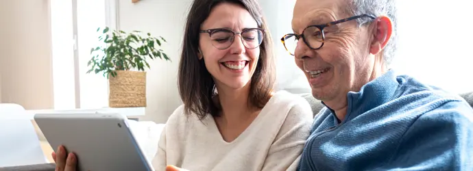 Eine Frau und ein älterer Mann lachen und teilen sich ein Tablet auf der Couch.