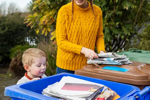 Ένα νεαρό αγόρι κοιτάζει μέσα σε έναν κάδο ανακύκλωσης γεμάτο με εφημερίδες, μια γυναίκα παρακολουθεί.