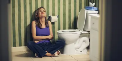Eine Frau hält ihren Bauch, während sie neben einer Toilette in einem gestreiften Badezimmer sitzt.