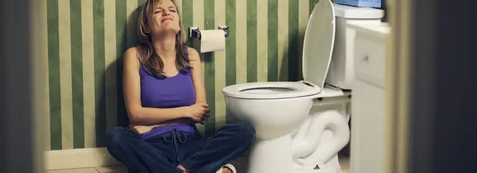 Eine Frau hält ihren Bauch, während sie neben einer Toilette in einem gestreiften Badezimmer sitzt.