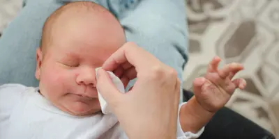 Beba zatvorenih očiju leži na nečijem krilu dok joj odrasla osoba nežno briše lice vlažnom maramicom.