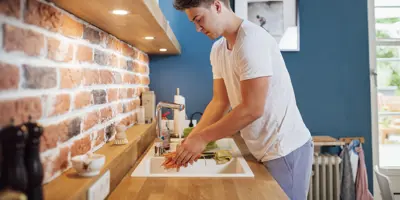 Ein Mann in einem weißen T-Shirt wäscht Möhren in einer Spüle vor einer Ziegelrückwand.