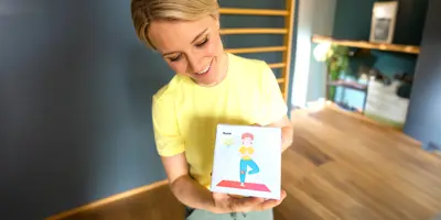 Lachende Frau in einem gelben Shirt hält eine Yogakarte mit einem Kind in Baumposition als Illustration in den Händen.