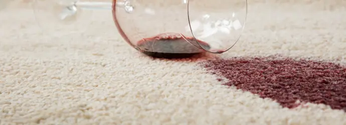 Ein umgekipptes Weinglas vergießt Rotwein auf einen beigen Teppich.
