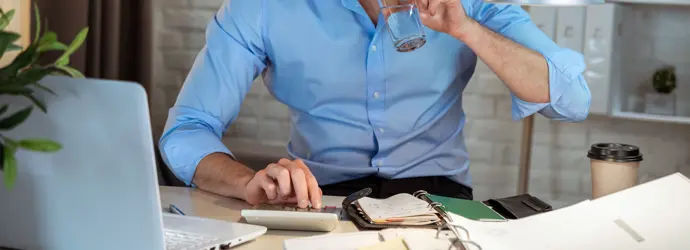 Ein Mann sitzt an einem Schreibtisch und trinkt ein Glas Wasser während er einen Taschenrechner benutzt.