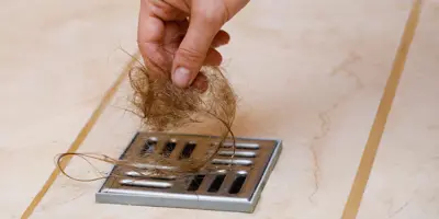 Eine ausgestreckte Hand zieht lange, hellbraune Haare aus einem Duschabfluss.