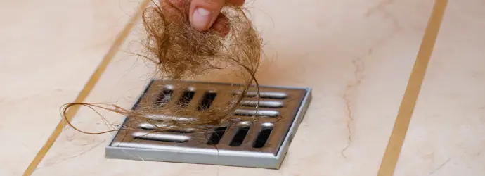 Eine ausgestreckte Hand zieht lange, hellbraune Haare aus einem Duschabfluss.
